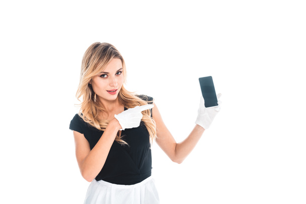blonde housemaid in black uniform and gloves pointing with finger on smartphone isolated on white - Foto, Bild