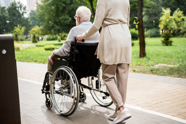 частковий вид на старшу жінку, яка носить чоловіка в інвалідному візку під час прогулянки в парку
 - Фото, зображення