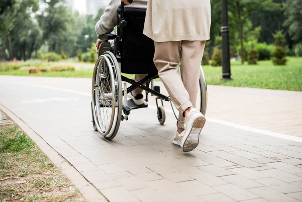 partial view of senior woman carrying husband in wheelchair while walking in park - 写真・画像