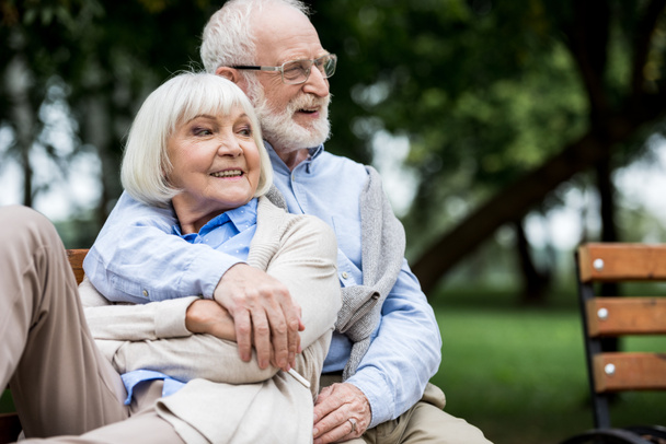 smiling senior man embracing happy wife while sitting together on bench in park - Fotoğraf, Görsel