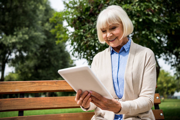 smiling senior woman using digital tablet while sitting on wooden bench in park - Photo, Image