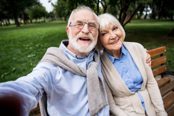 selective focus of happy smiling senior couple sitting on wooden bench in park - Φωτογραφία, εικόνα