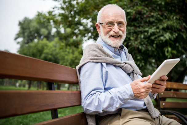 valikoiva painopiste hymyilevä vanhempi mies käyttää digitaalinen tabletti istuessaan penkillä puistossa
 - Valokuva, kuva