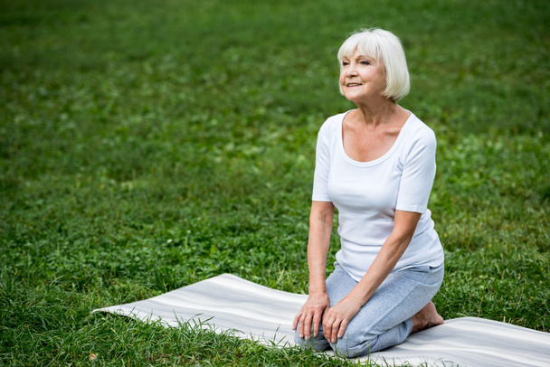 mosolyogva ül a jóga szőnyeg, meditációs póz, kezével térdét idősebb nő - Fotó, kép