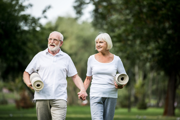 Lächelndes Senioren-Paar mit Fitnessmatten, das im Park spaziert und Händchen hält - Foto, Bild