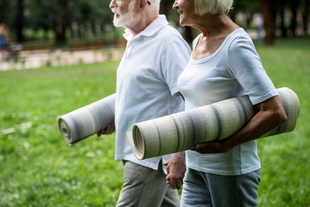 vista parcial de la sonriente pareja de ancianos con colchonetas de fitness caminando por el parque y tomados de la mano
 - Foto, imagen