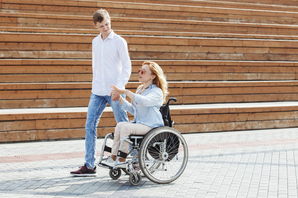 Женщина в инвалидной коляске с бойфрендом прогуливается в городском летнем парке
 - Фото, изображение