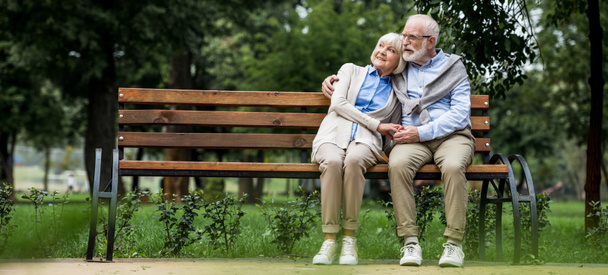 gelukkige senior paar knuffelen terwijl zittend op houten bankje in park - Foto, afbeelding