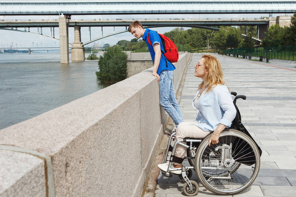 Жінка в інвалідному візку з хлопцем, що гуляє в міському літньому парку
 - Фото, зображення