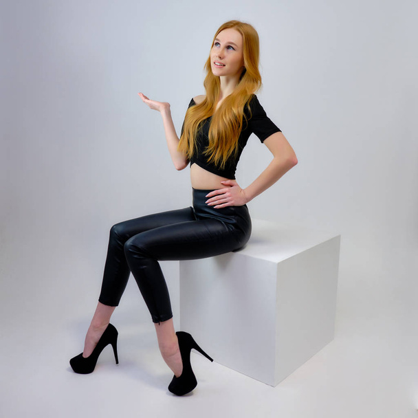 Concept portrait of a pretty beautiful red-haired girl talking on a gray background in the studio sitting on a cube in front of the camera. They show different emotions in different poses. - Фото, изображение