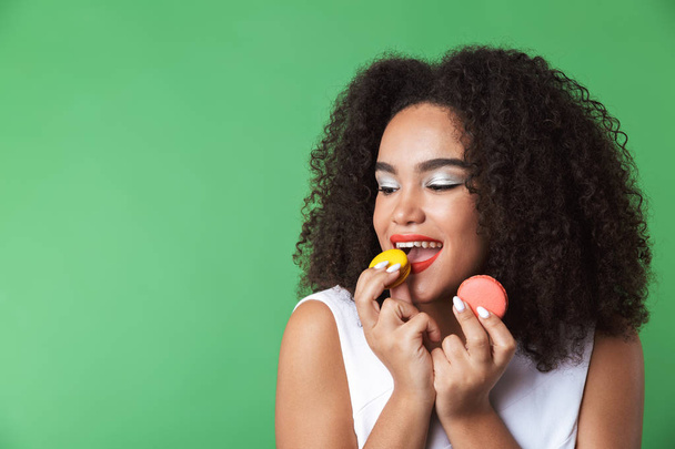 Cheerful young african woman wearing dress standing isolated, showing colorful macaroons - Фото, изображение