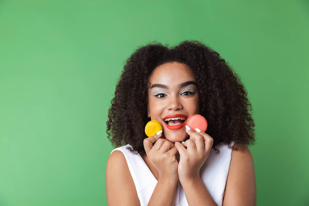 Cheerful young african woman wearing dress standing isolated, showing colorful macaroons - Foto, Bild