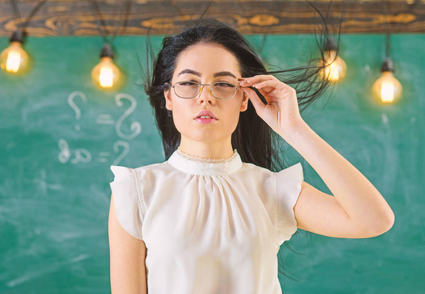 Γυναίκα με μακριά μαλλιά με λευκή μπλούζα στέκεται στην τάξη. Κυρία αυστηρός δάσκαλος στο ήρεμο πρόσωπό στέκεται μπροστά από το μαυροπίνακα. Έννοια του αυστηρή δασκάλα. Δασκάλα με γυαλιά και κυματίζοντας τα μαλλιά φαίνεται ελκυστικό - Φωτογραφία, εικόνα