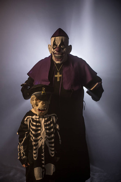 Dark priest in scary clown mask and skeleton boy posing with money and gun against grey background - Foto, imagen