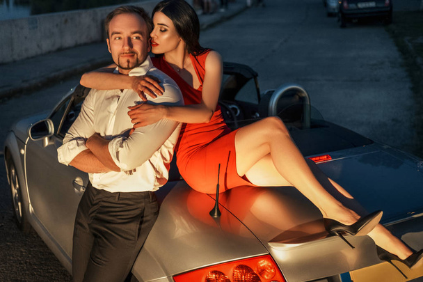 Gorgeous sexy copule of man and woman on a vacation car trip in summer day. Luxury grey sport car. Sexy fit brunette woman and man in evening outfit. Night portrait. - Valokuva, kuva