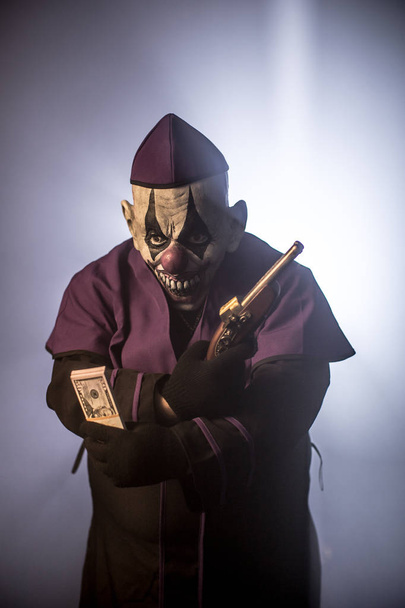 Dark priest in scary clown mask posing with money and gun against grey background - Photo, image