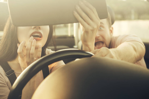 Gorgeous sexy copule of man and woman on a vacation car trip in summer day. Husband screams until his wife applying lipstick using the rear view mirror in the moving car. Distracted dangerous driving. - Photo, Image