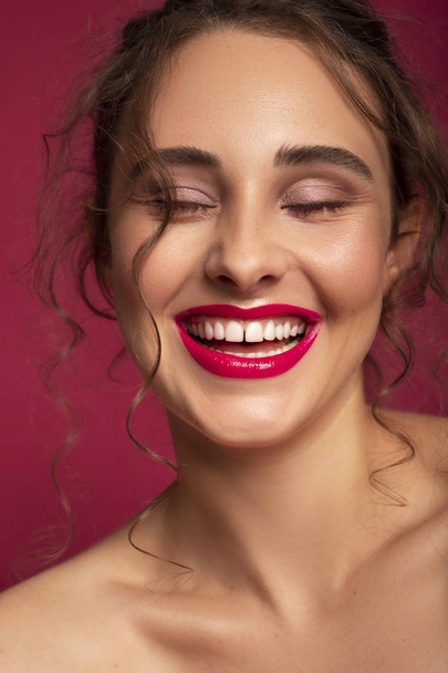 Nettes brünettes Model mit makelloser Haut und kreativem buntem Make-up auf dunkelrosa Hintergrund. Sie trägt einen lustigen Dutt als Frisur und lässt sich solide rosa Lippen schminken. - Foto, Bild