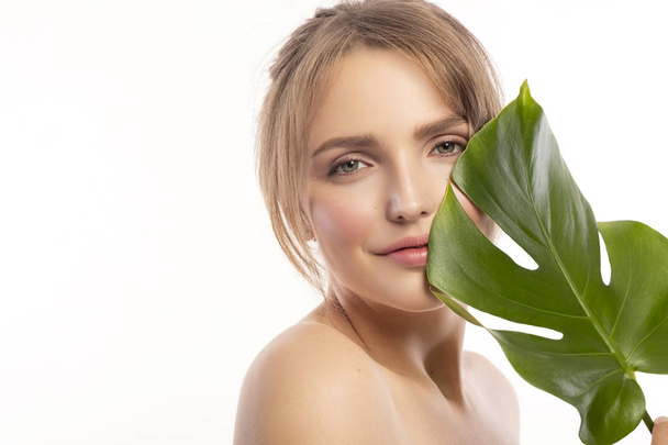 Cute smiling blonde caucasian woman with fresh, clean, healthy, flawless skin. Nude, day make up with casual hairstyle. Portrait on a white background with tropical monstera leaf near model face. - Photo, Image
