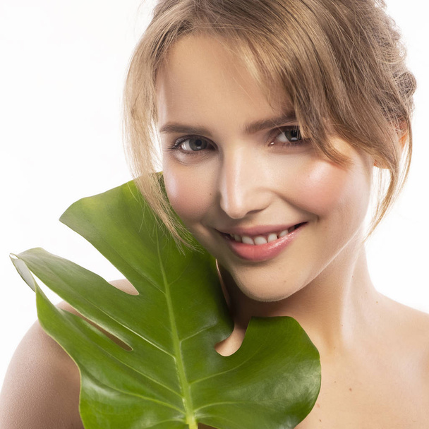 Cute smiling blonde caucasian woman with fresh, clean, healthy, flawless skin. Nude, day make up with casual hairstyle. Portrait on a white background with tropical monstera leaf near model face. - Photo, Image