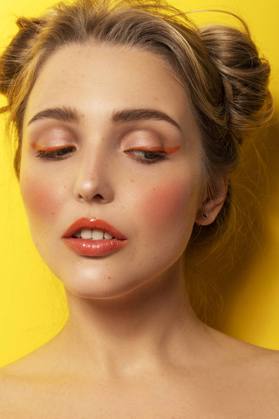 Nettes blondes Model mit makelloser Haut und kreativem buntem Make-up auf leuchtend gelbem Hintergrund. sie trägt 2 lustige Brötchen als Frisur. - Foto, Bild