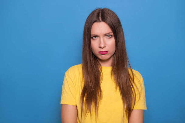 Cute brunette woman with long hair posing in yellow t-shirt on a blue background. Emotional portrait. She sad, cry - Photo, image