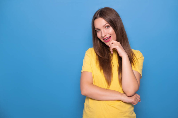Nette brünette Frau mit langen Haaren posiert in gelbem T-Shirt auf blauem Hintergrund. Emotionales Porträt. sie lächelt glücklich mit makellosen weißen Zähnen und flirtet - Foto, Bild