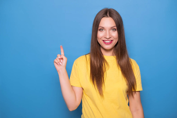 Cute brunette woman with long hair posing in yellow t-shirt on a blue background. Emotional portrait. She smiles happily with flawless white teeth and poins finger on something - Foto, immagini