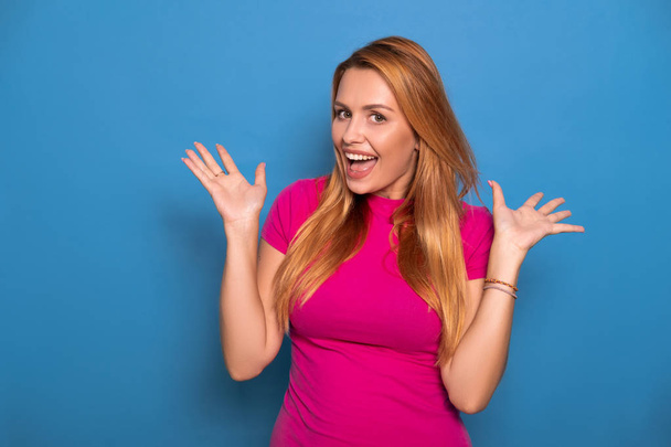 Sexy plus size model with long red hair in pink t-shirt on a blue background. Emotional portrait. She throw her arms up in amazement, smiling - Valokuva, kuva