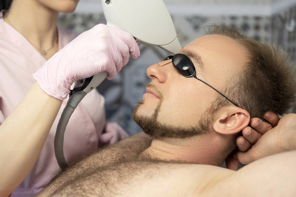 Woman doctor cosmetologist removing hair from a man body using laser epilator. They in a spa center. - Photo, Image