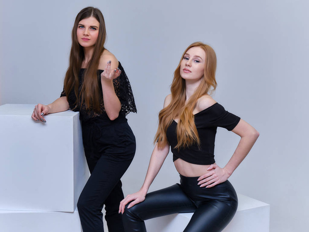Concept portrait of two cute beautiful girls talking on a gray background in the studio sitting on the cubes in front of the camera. They show different emotions in different poses. - Фото, зображення