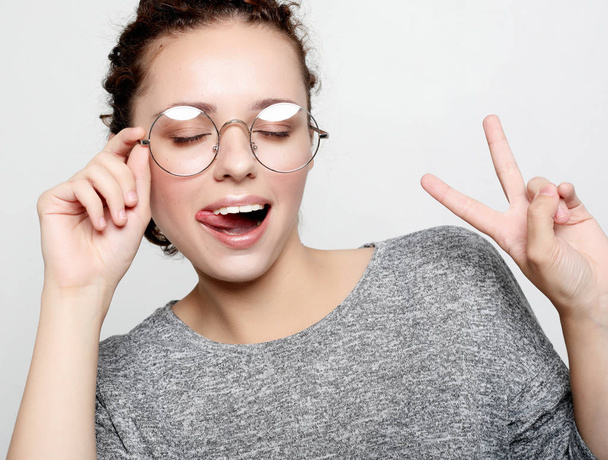 Jovem mulher encaracolada vestindo óculos vestidos casualmente fazendo rostos na câmera, piscando, salientando a língua
. - Foto, Imagem