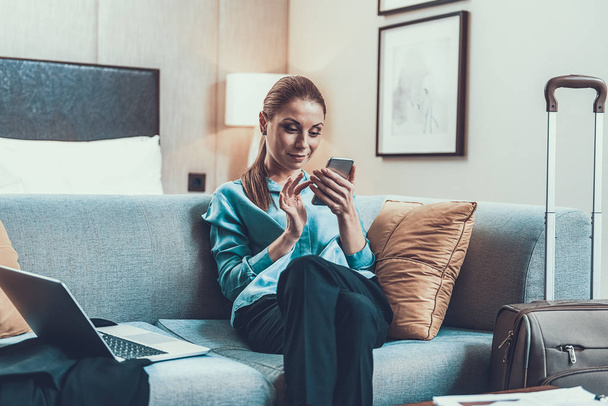 Elegant lady sitting on the sofa in hotel room and using smartphone - Фото, изображение
