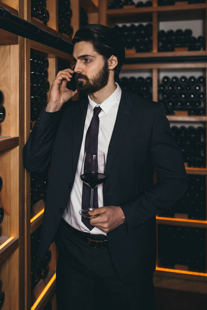 Молодой винодел нюхает аромат красного вина в стакане, он проверяет цвет, качество и отложения напитка
.  - Фото, изображение