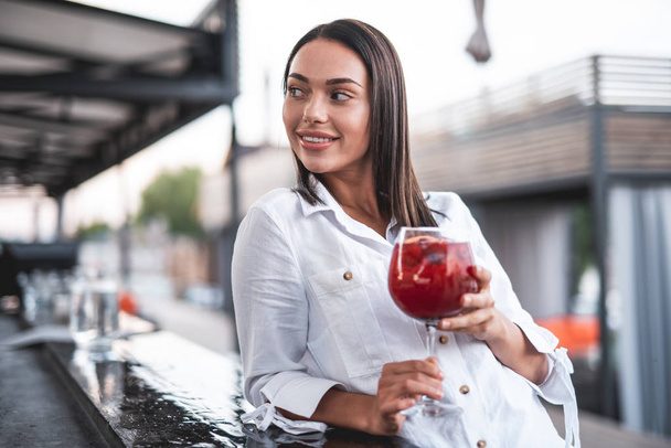 Waist up of relaxed young lady putting one elbow on the bar counter and holding her cocktail while smiling and looking away - Фото, зображення