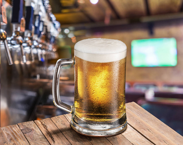 Ayaz kupa eski ahşap masa üzerinde hafif bira. Pub iç ve bar counter bira ile arka plan musluklar. - Fotoğraf, Görsel