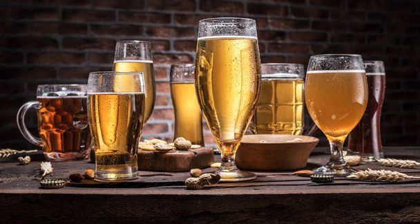 Cold mugs and glasses of beer on the old wooden table, brick wall at the background. Assortment of beer. - Fotografie, Obrázek