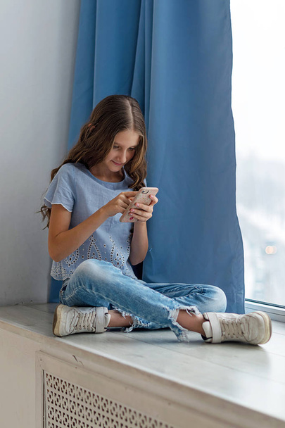 Portrait of a Caucasian teenage girl sitting near the window with a mobile phone. Educational concept. Modern youth. - Photo, image
