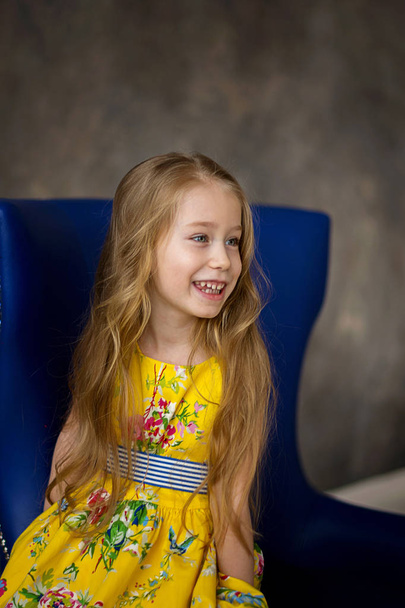 Retrato de una niña alegre con un vestido amarillo brillante en una silla azul. Brillante color. Tendencia. Modelo pequeño
. - Foto, Imagen