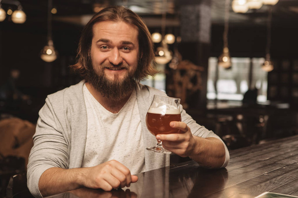 Cheerful young bearded man smiling happily to the camera, holding out glass of delicious craft beer, copy space. Happy man enjoying tasty beer at local pub. Excited male client resting at the bar - Foto, Imagen
