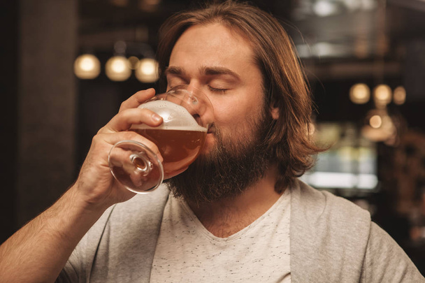 Close up of a bearded man sipping delicious craft beer with his eyes closed, resting at the bar. Happy relaxed male customer drinking beer, celebrating holidays at the pub. Oktoberfest, relax concept - Photo, image