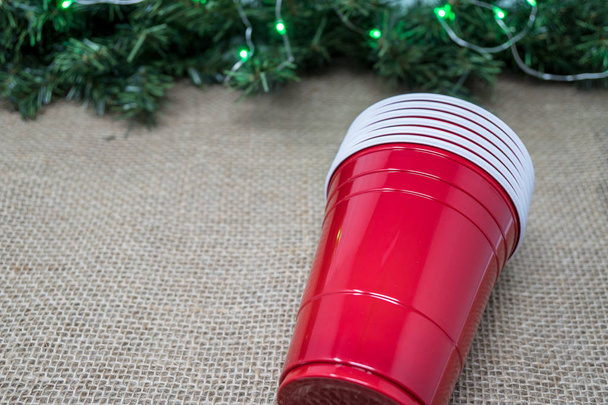 Stapel roter Partybecher isoliert auf einem Plateau-Hintergrund mit Weihnachtsgirlanden. Konzept für eine Weihnachtsfeier, bei der Alkohol ausgeschenkt wird - Foto, Bild