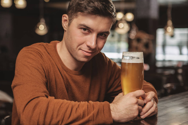 Cheerful attractive man smiling confidently to the camera, relaxing with a glass of beer at the counter of a beer pub. Handsome man enjoying delicious craft beer at the bar, copy space - Φωτογραφία, εικόνα