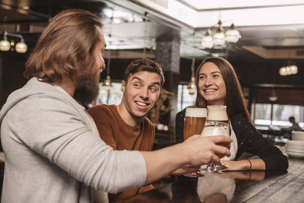Handsome young man smiling happily, enjoying resting at the bar with his friends. Group of friends having drinks at the local beer pub. Two men and beautiful woman drinking beer. Celebration concept - Photo, Image