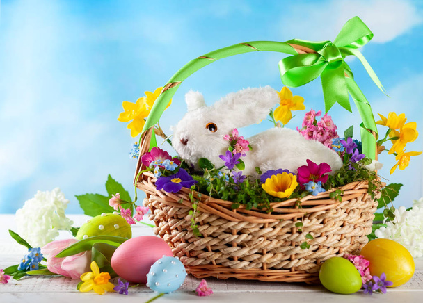Osterkomposition mit kleinen weißen Hasen im Korb, Frühlingsblumen und bunten Ostereiern. Osterkarte mit Kopierraum. - Foto, Bild