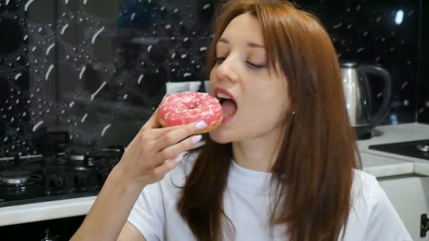 自宅のキッチンで赤いドーナツを食べて空腹若い髪の長い女。おいしい食品のコンセプト - 映像、動画