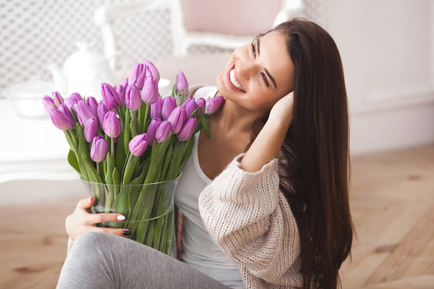 Красивая женщина в постели держит цветы и читает записку. Молодая девушка улыбается. Веселая женщина с тюльпанами
 - Фото, изображение