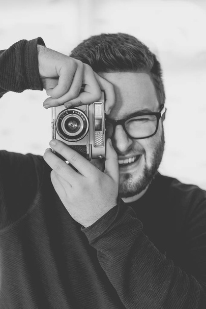 Zblízka, černobílý portrét hezký, mladý muži, nosí brýle a fotografování s vintage, 35mm fotoaparát zatímco usmívá na kameru - Fotografie, Obrázek