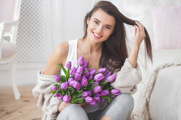 Attractive young woman with flowers indoors in the bedroom. Portrait of beautiful lady at home. Close up shot of female with tulips. - Valokuva, kuva
