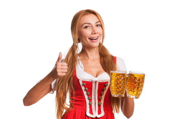Beautiful happy Bavarian waitress smiling cheerfully to the camera showing thumbs up while holding two mugs of delicious beer isolated on white, copy space on the side. Oktoberfest, drinking, party - Fotoğraf, Görsel